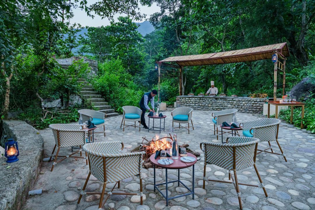 An outdoor dining area with wicker chairs and a fire pit, surrounded by stone steps and greenery.