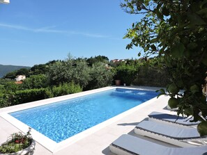 Ein rechteckiger Pool mit klarem, blauem Wasser, umgeben von einer weißen Fliesenveranda. Entlang des Poolrandes stehen weiße Liegestühle mit schwarzen Kissen.