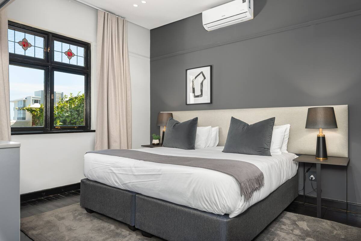 A modern bedroom with a large bed, two bedside tables, a window with curtains, and a wall-mounted air conditioner.