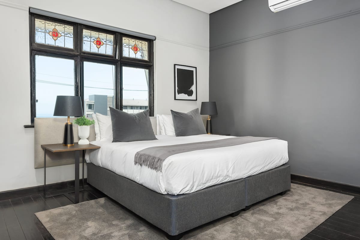 A modern bedroom with a large bed, two bedside tables, a potted plant, and a window with stained glass.