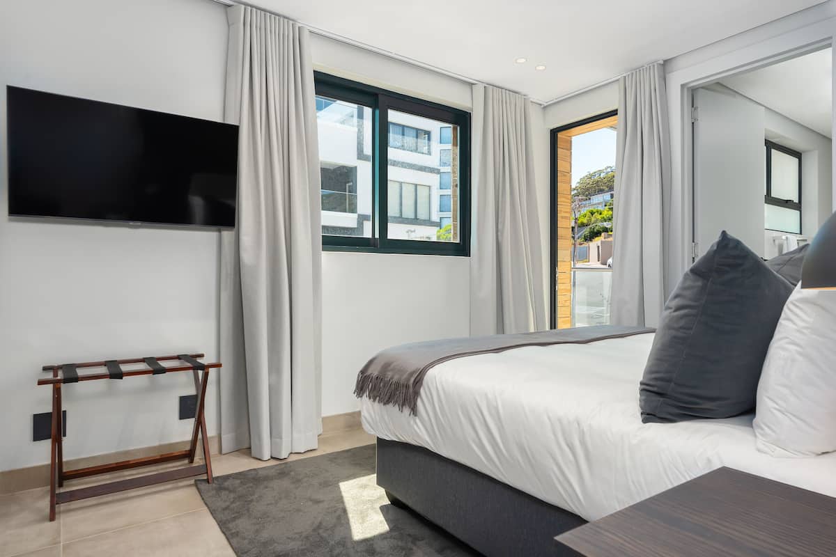 A modern bedroom with a large bed, a TV mounted on the wall, and a view of the outside through a window.