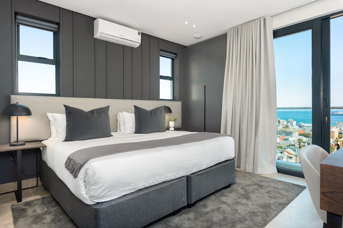 A modern bedroom with a large bed, grey bedding, and a city view through a large window.