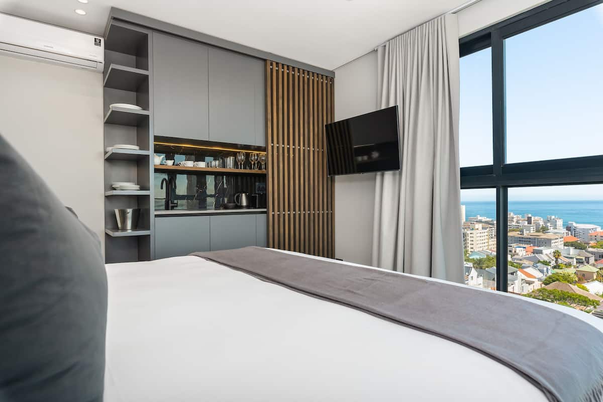 A modern hotel room with a large bed, a built-in kitchen area, a TV, and a city view.