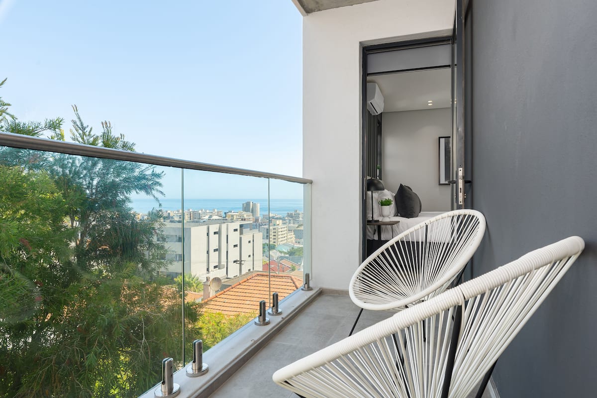 A balcony with a view of the city and sea, white wicker chairs, and a small shelf with a plant.