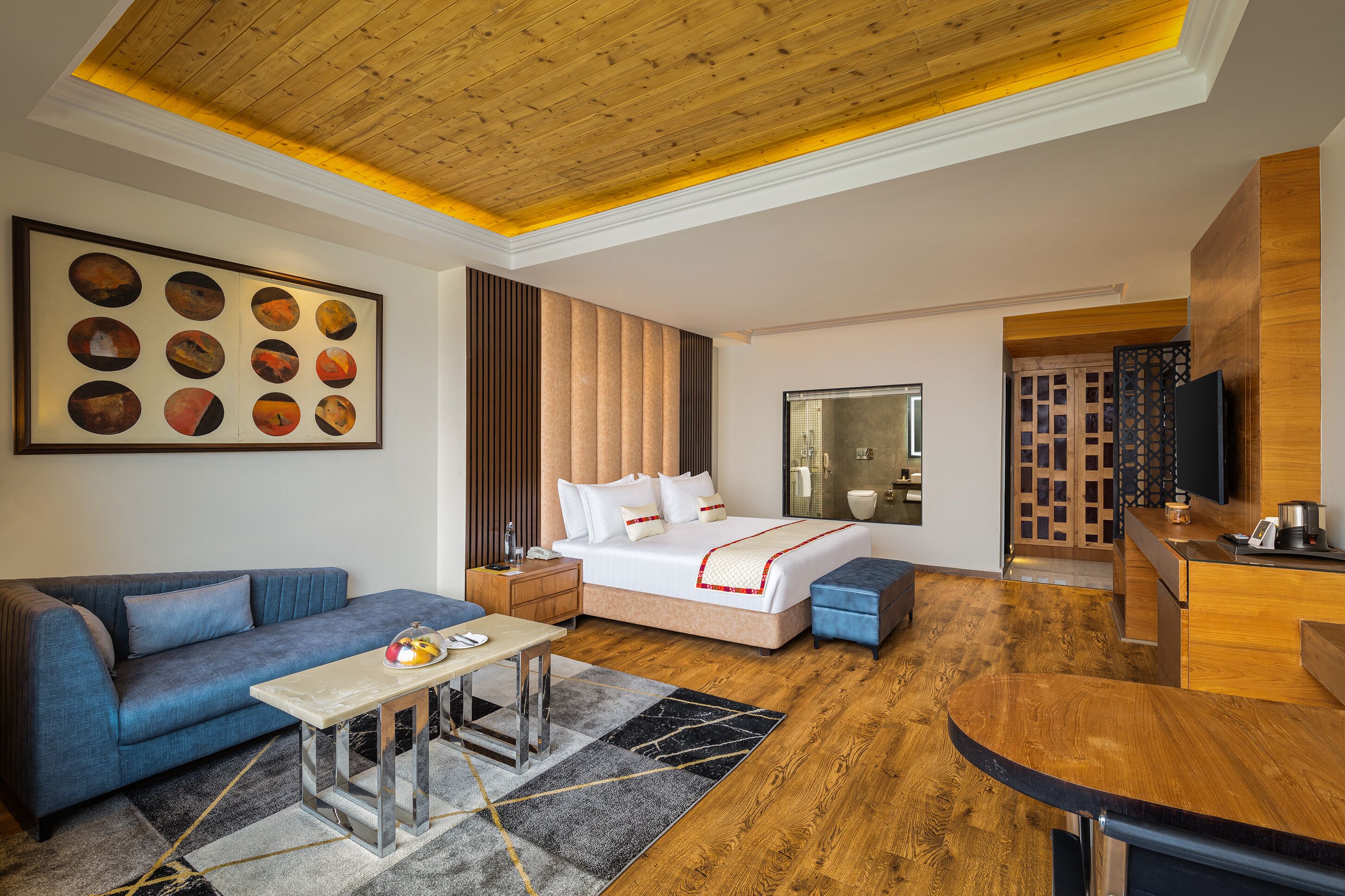 Una habitación de hotel moderna con techo de madera, una cama grande con ropa blanca, un sofá azul, una mesita pequeña y un escritorio de madera.