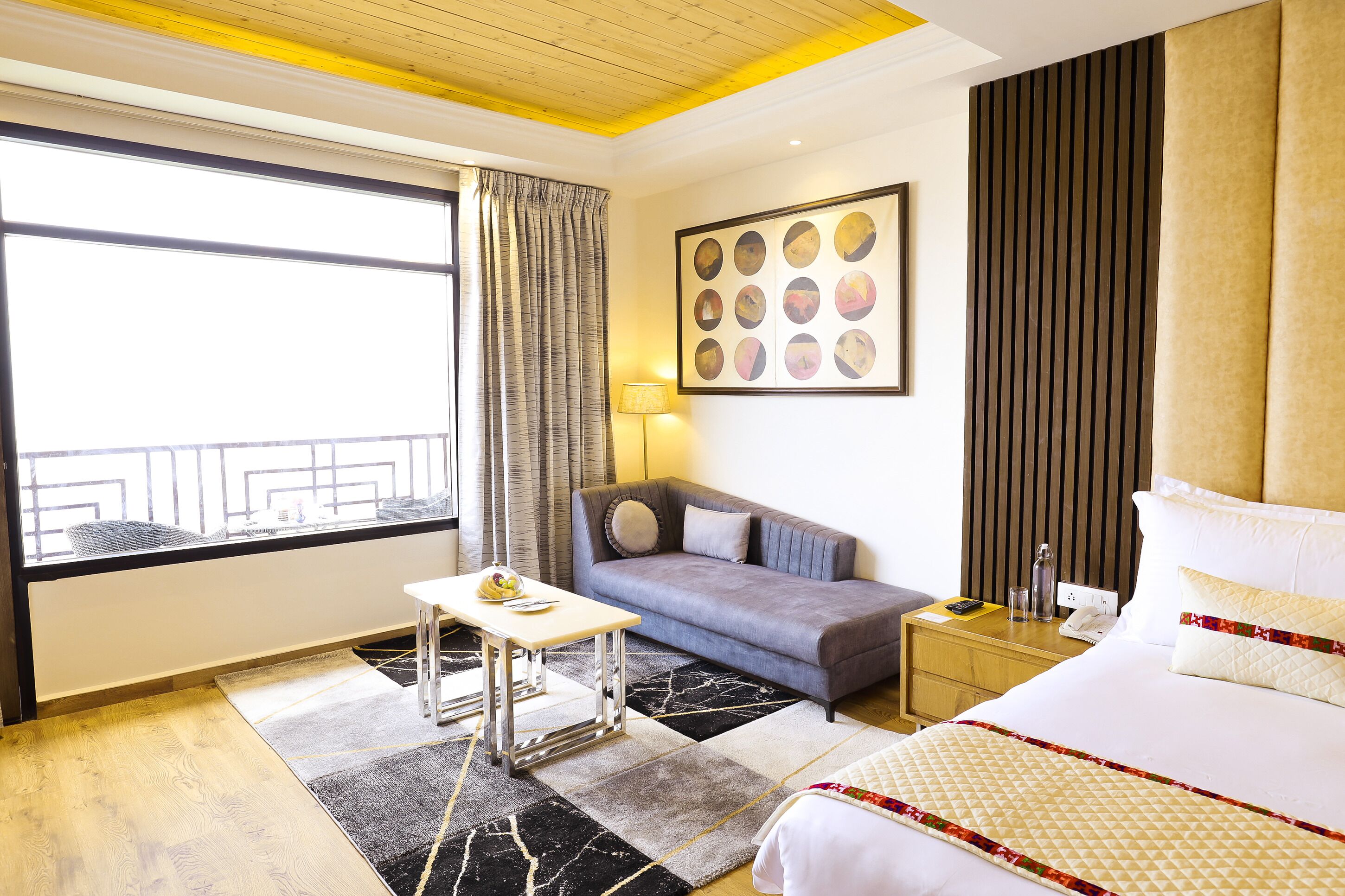 A hotel room with a bed, a sofa, a small table, a lamp, and a large window with curtains.