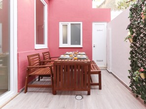 A wooden table set for breakfast with plates, glasses, and orange juice, surrounded by chairs, against a pink wall with a window and a door.