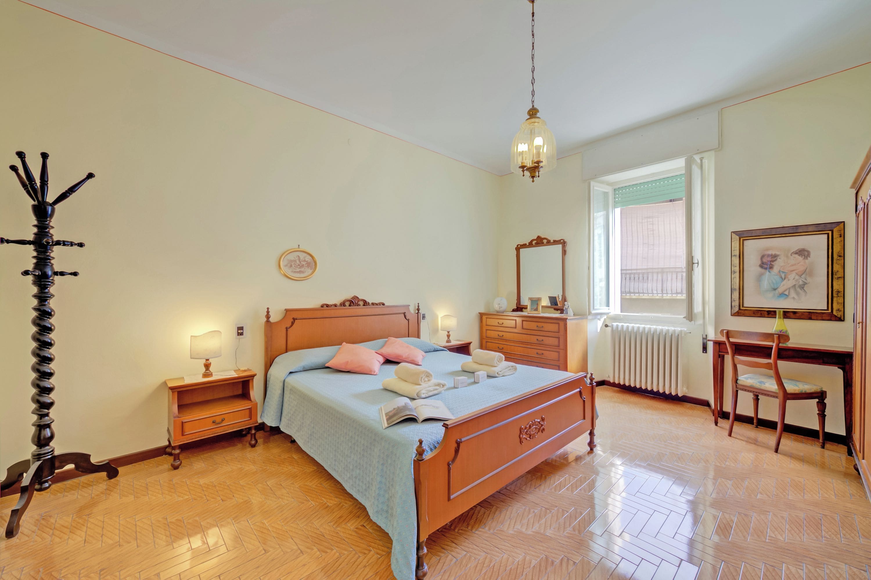 A bedroom with a wooden bed, bedside tables, a chair, a desk, a radiator, and a window with blinds.
