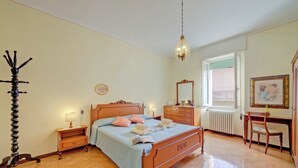 Une chambre à coucher avec un lit en bois, des tables de chevet, une chaise, un bureau, un radiateur et une fenêtre avec des stores.