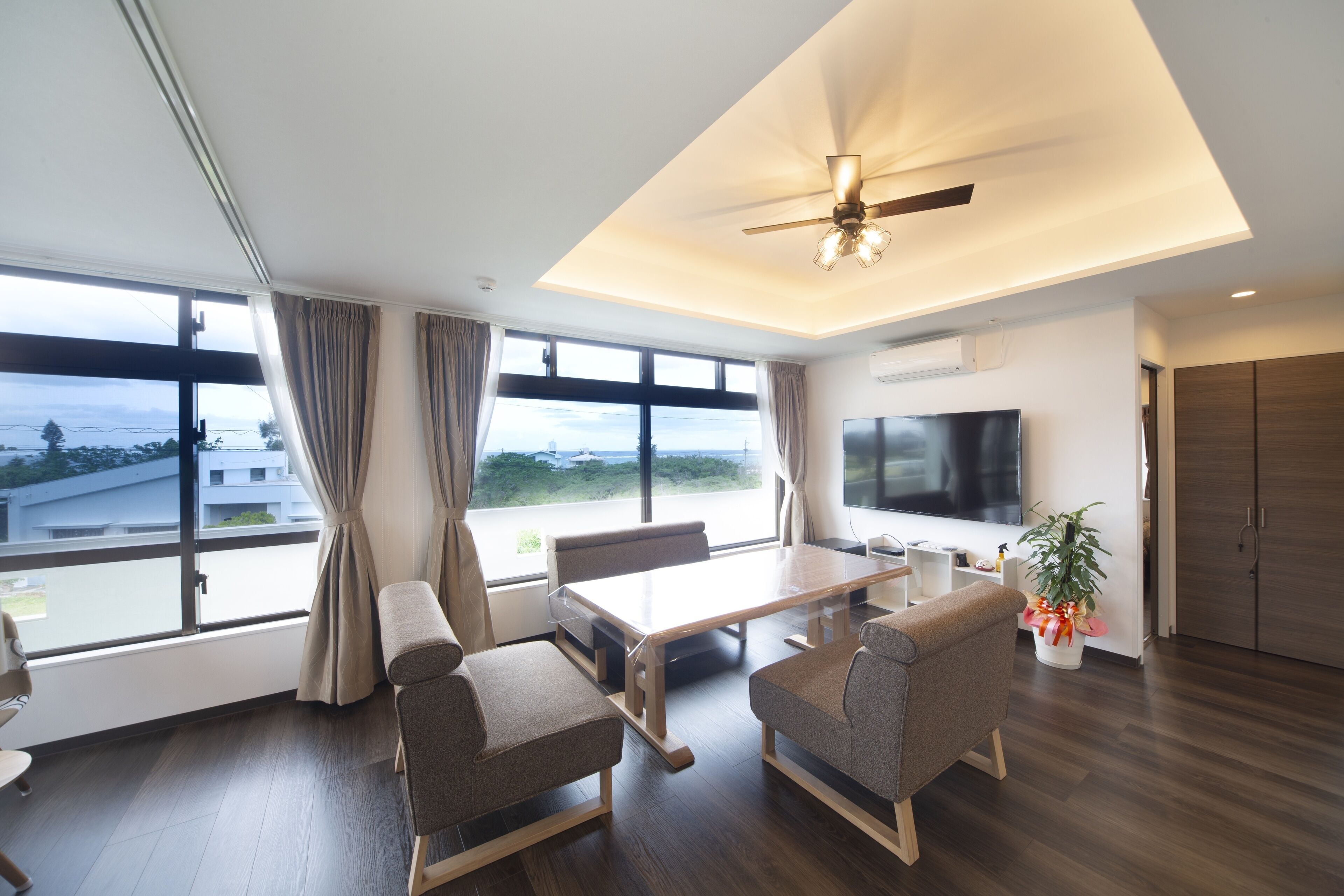 A modern living room with a large window, a ceiling fan, a flat-screen TV, and a dining area.