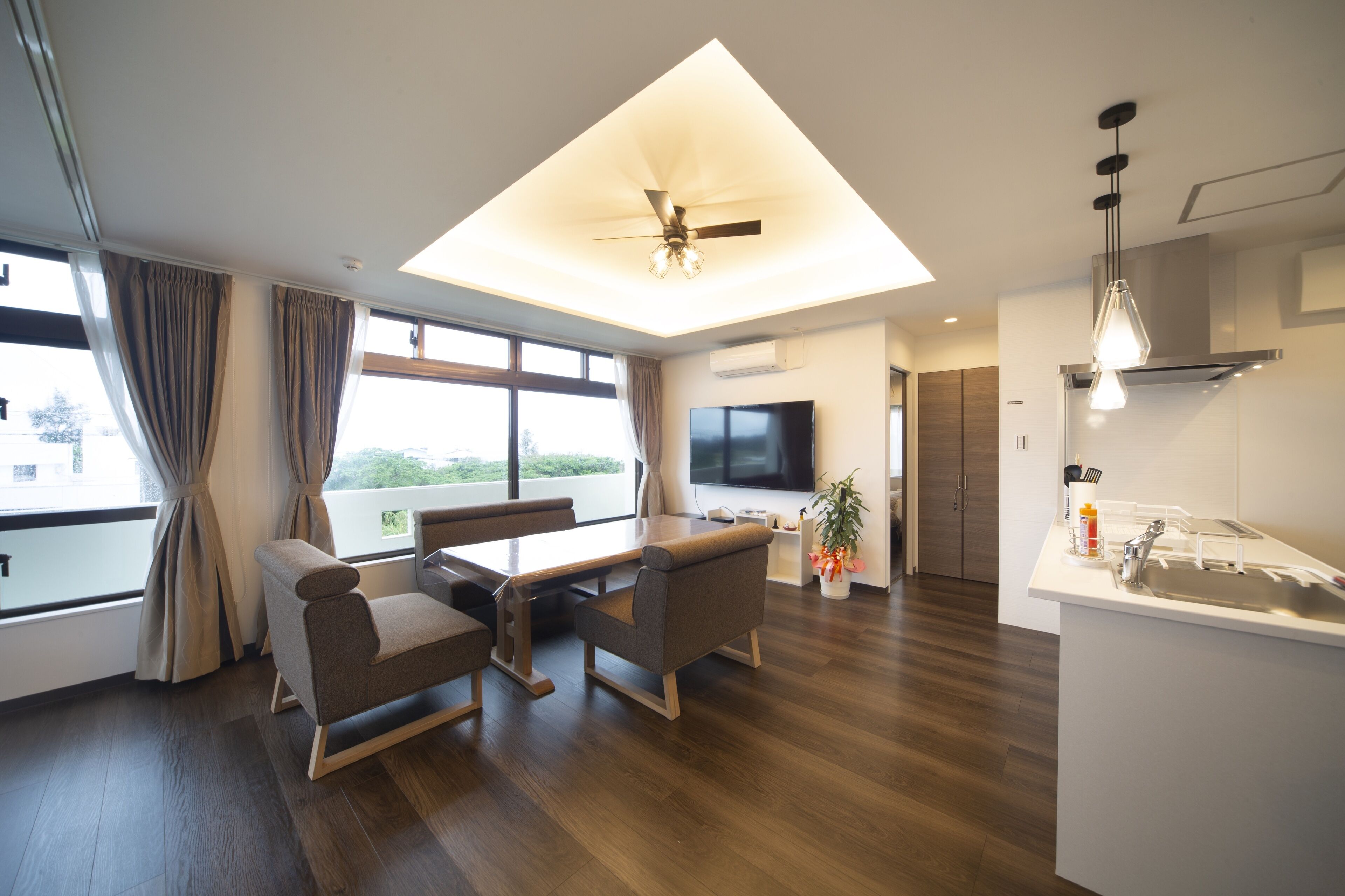A modern living room with a dining area, a ceiling fan, a flat-screen TV, and a kitchen with a sink and countertop.