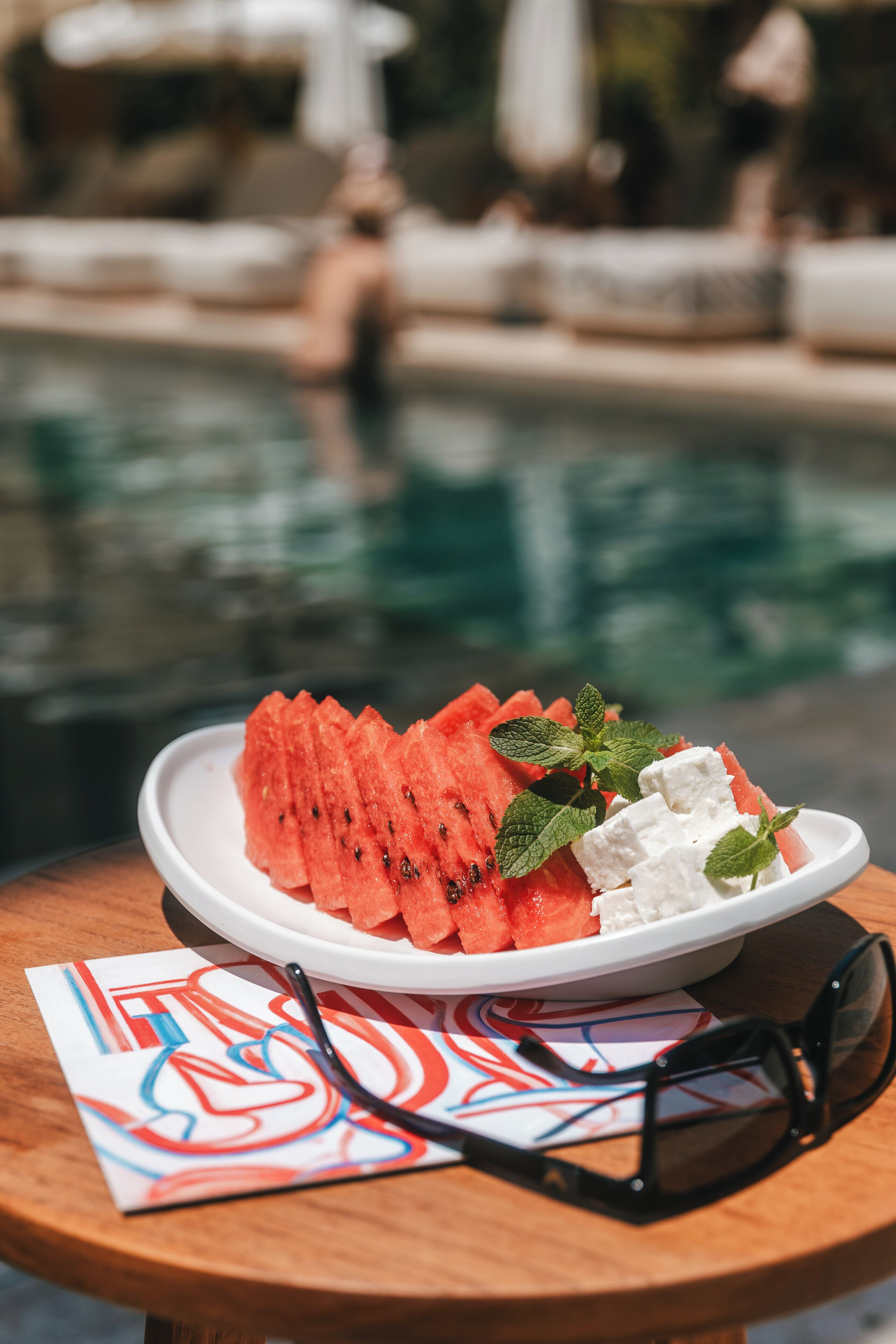 A plate of watermelon and feta cheese on a wooden table with a pair of sunglasses and a colorful napkin.