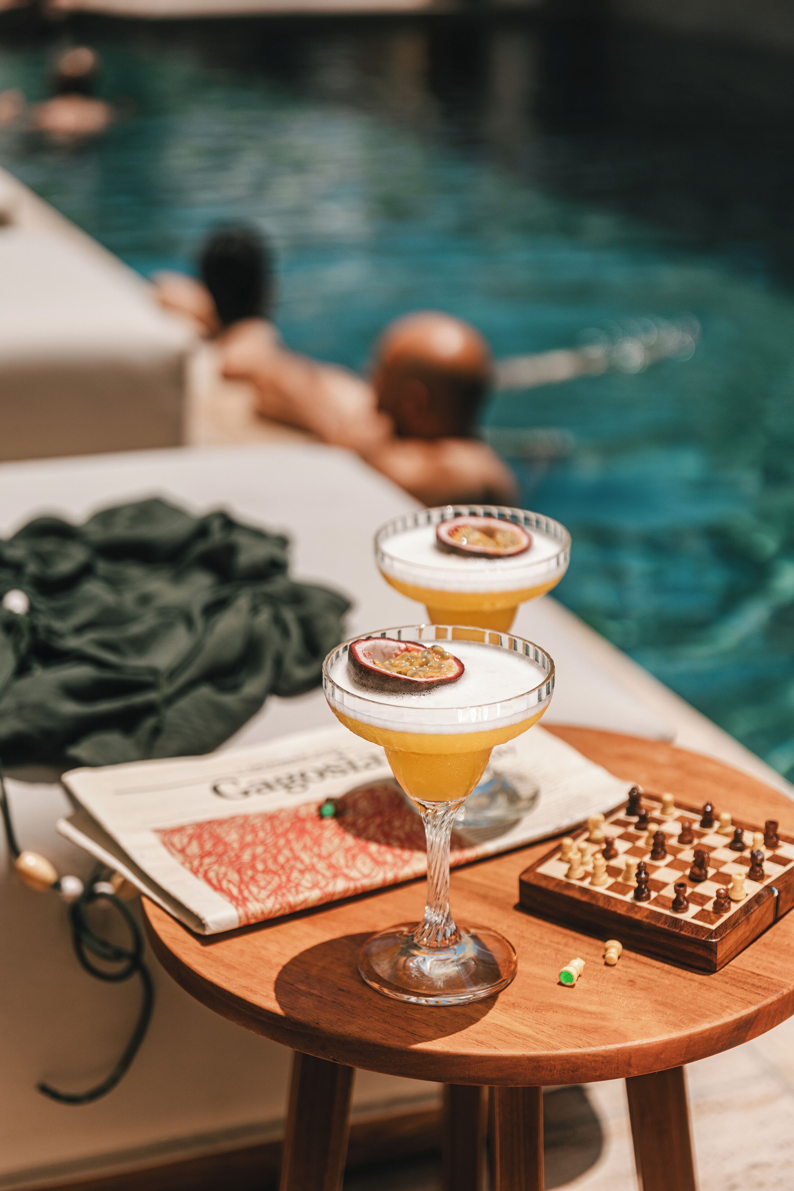 Two cocktails on a wooden table by a poolside, with a chessboard and a menu nearby.