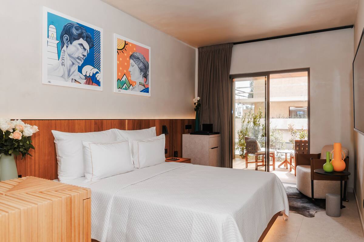 A hotel room with a large bed, a wooden headboard, a desk, a chair, a TV, and a balcony with outdoor furniture.
