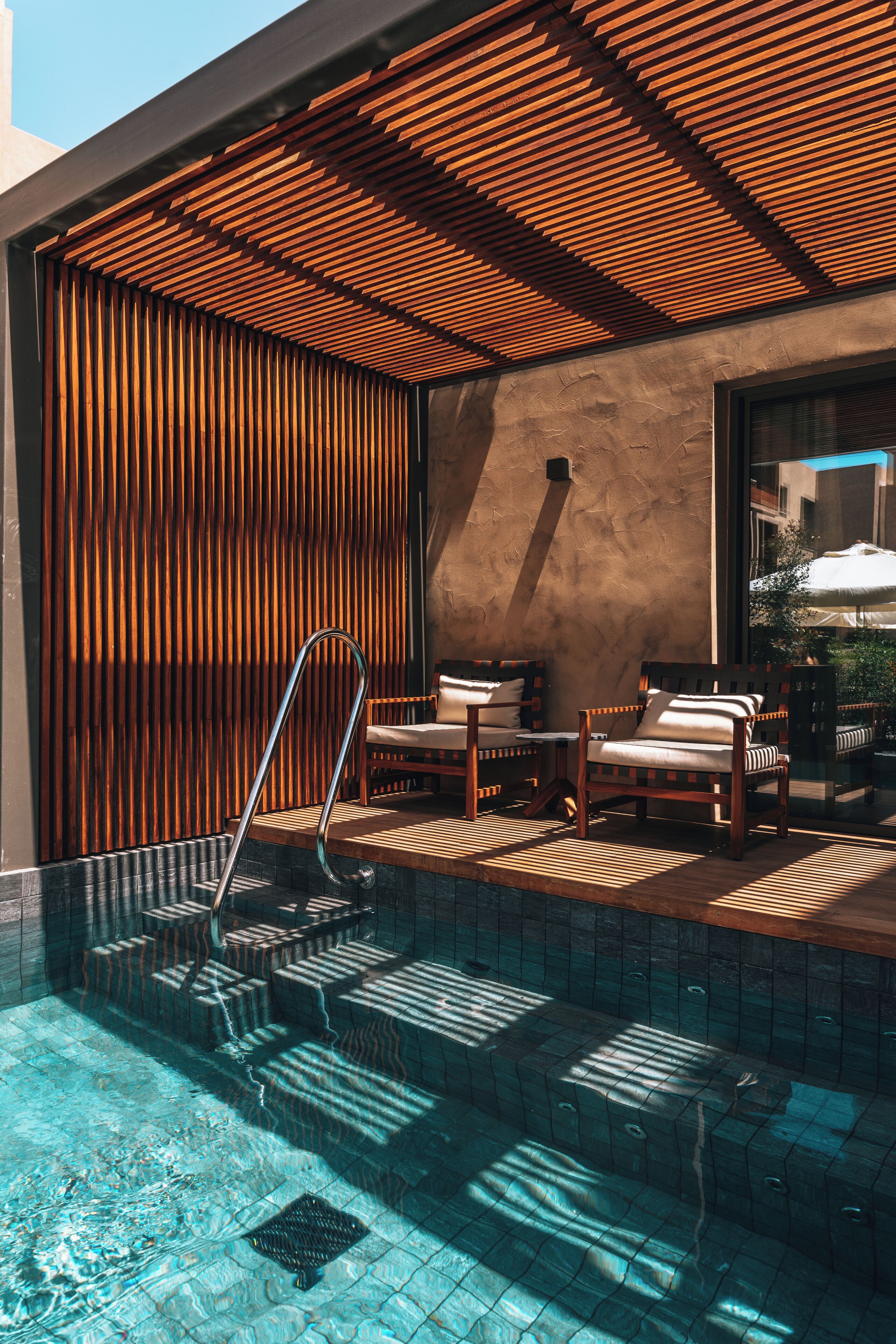 A pool area with wooden slatted ceiling, two lounge chairs, and a glass door leading outside.