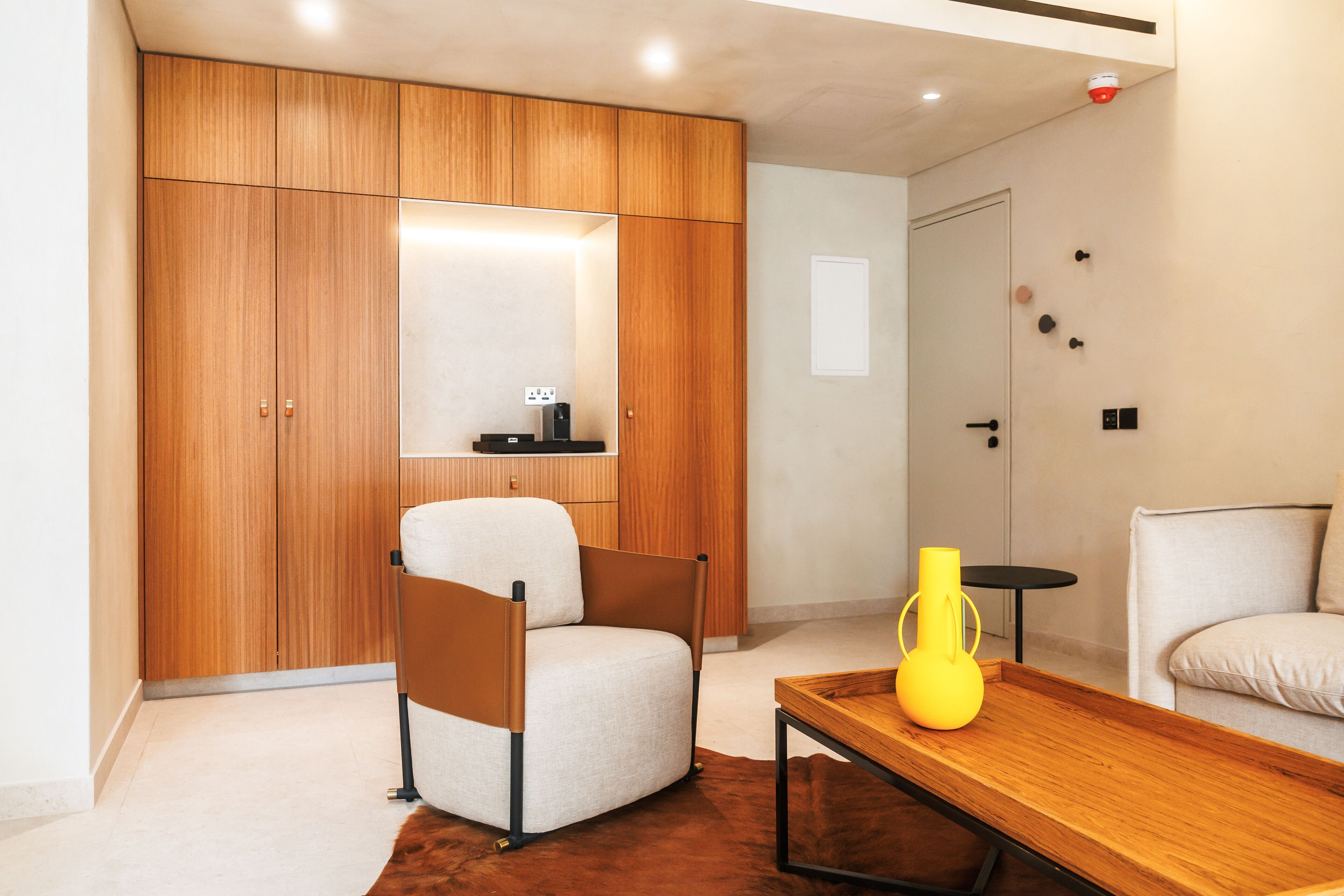 A modern room with a wooden wardrobe, a white armchair, a small table with a yellow vase, and a sofa.