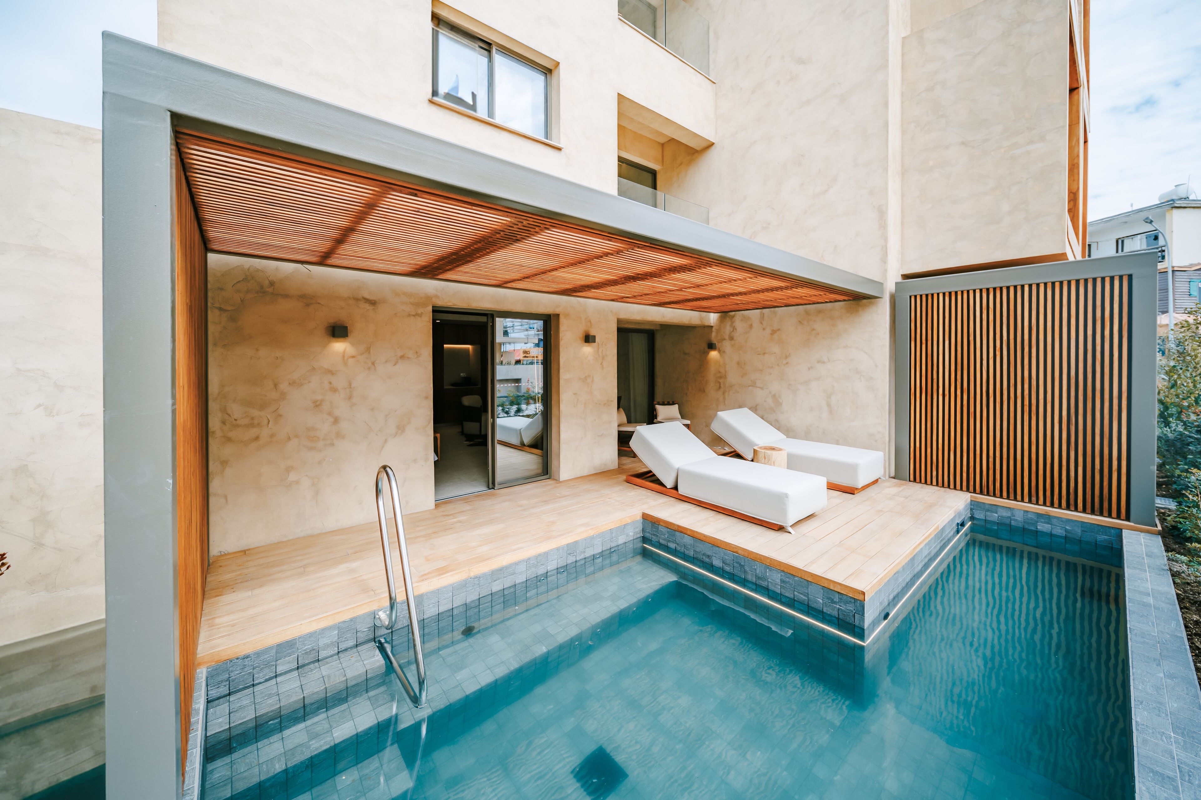 Un área de piscina moderna al aire libre con una terraza de madera, dos sillas de descanso blancas y un cuarto de spa con paredes de vidrio.