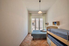 A small, well-lit room with a bunk bed, a single bed, a hanging light fixture, and a view through a sliding glass door.