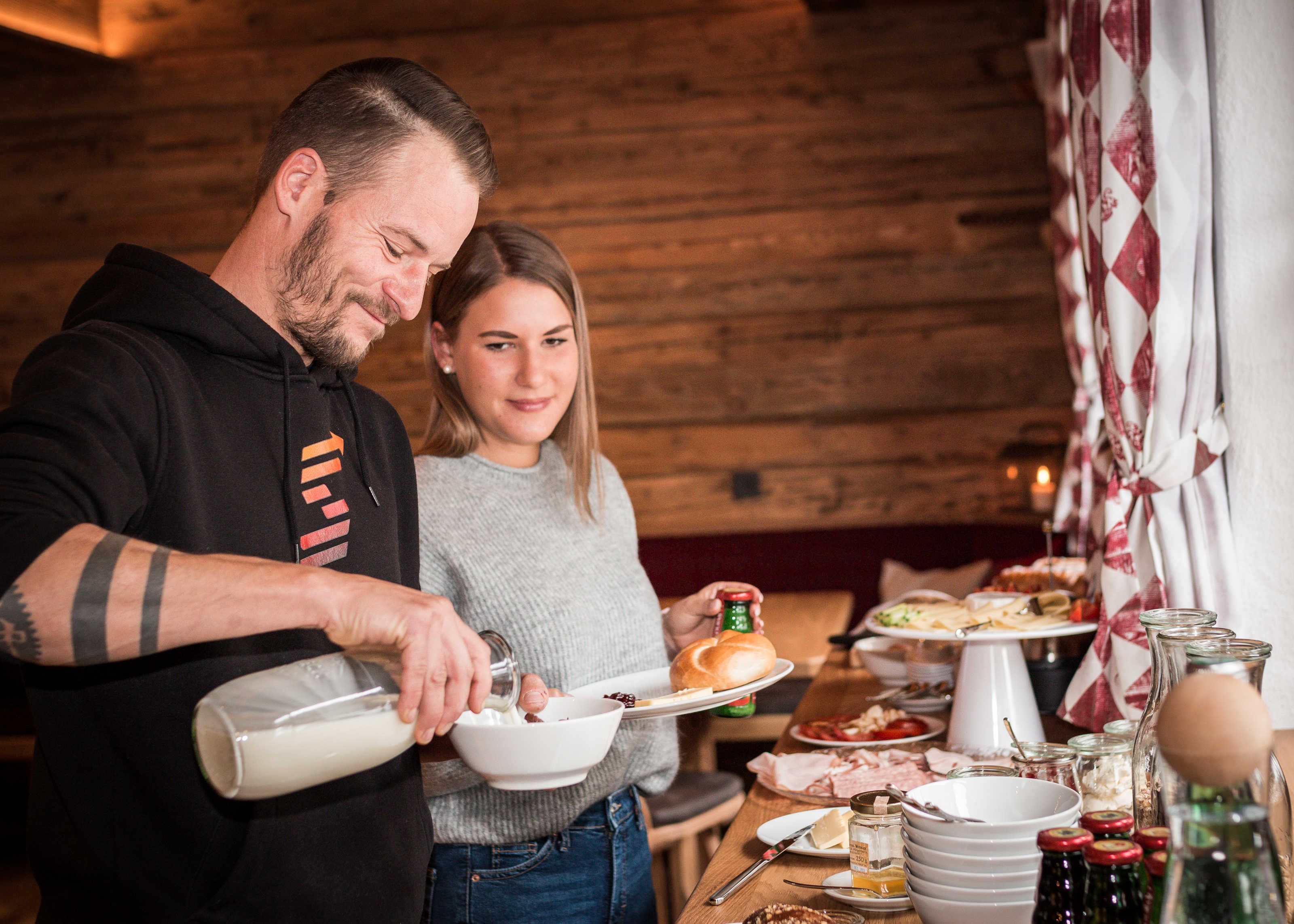 Un homme et une femme préparent des aliments dans un cadre rustique, avec des murs en bois et une table sur laquelle sont disposés divers plats et condiments.