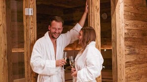 Un sauna en bois avec une porte en verre, une bouteille de champagne et deux personnes vêtues de robes blanches.