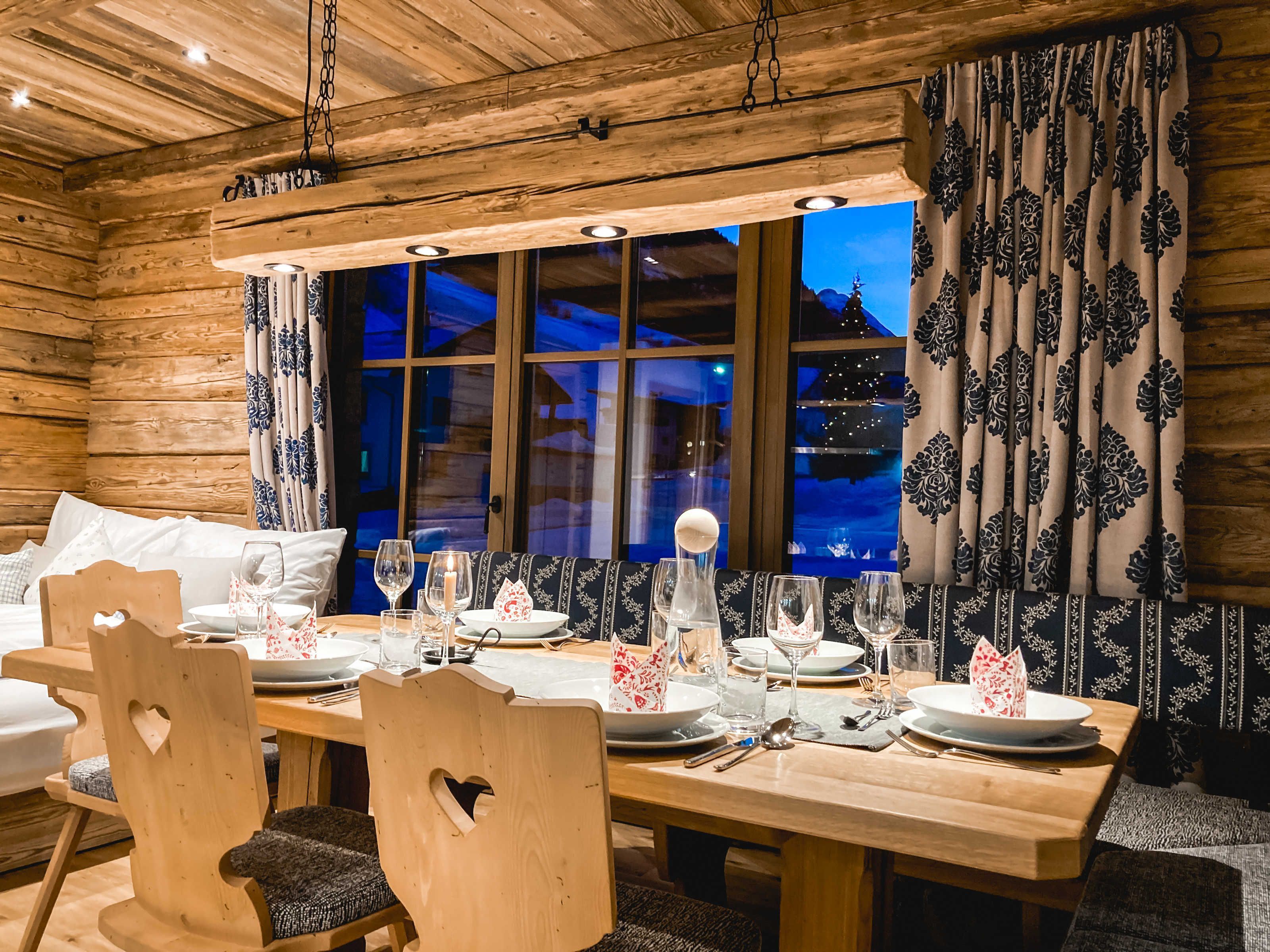 A wooden cabin-style room with a dining table set for four, wooden chairs, a large window with curtains, and a hanging light fixture.