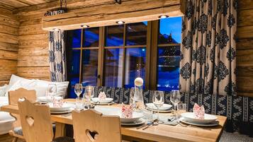 A wooden cabin-style room with a dining table set for four, wooden chairs, a large window with curtains, and a hanging light fixture.