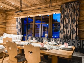 A wooden cabin-style room with a dining table set for four, wooden chairs, a large window with curtains, and a hanging light fixture. - Stadl-suite Silvretta - Stadl Chalet Ischgl (Ischgl)