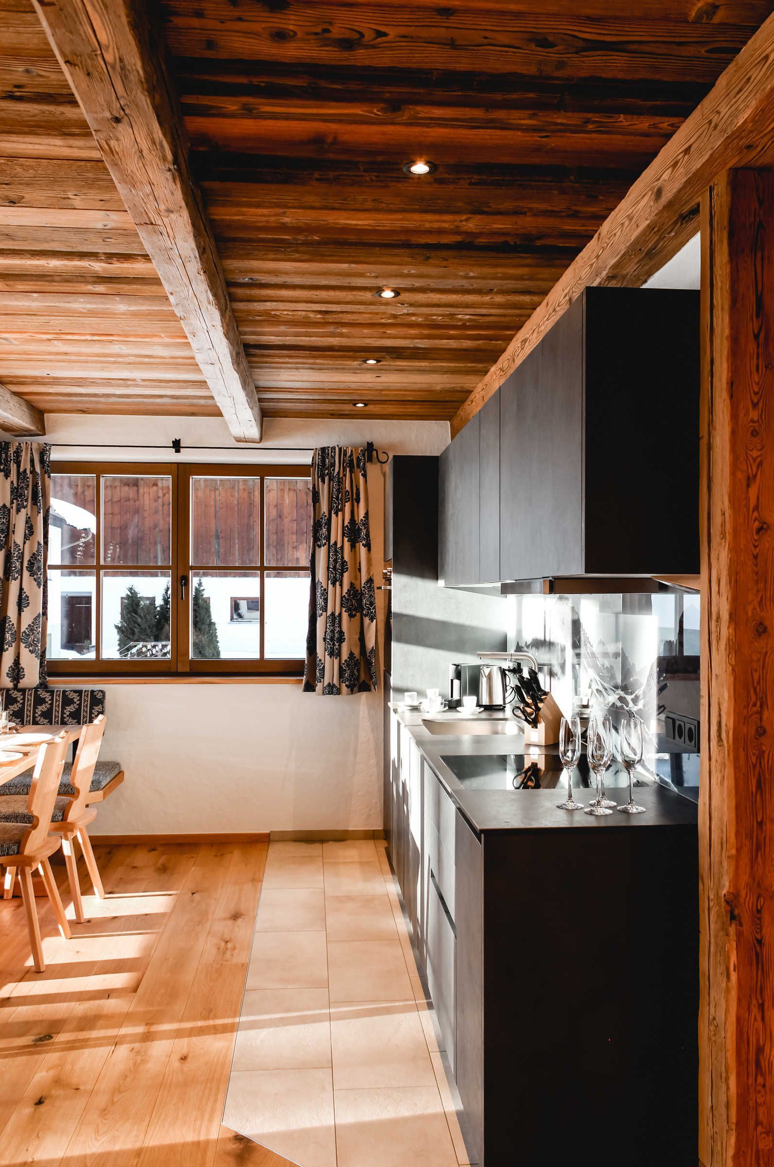 Eine moderne Küche mit Holzbalkendecke, einem Essbereich mit Stühlen und einem großen Fenster mit Vorhängen.