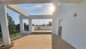 Un espacio al aire libre techado, con suelo de ladrillo, paredes blancas y vistas a árboles y cielo.