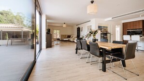 Un amplio comedor con una mesa de madera y sillas modernas, un ventanal con vistas al exterior y una zona de cocina con armarios oscuros y electrodomésticos blancos.