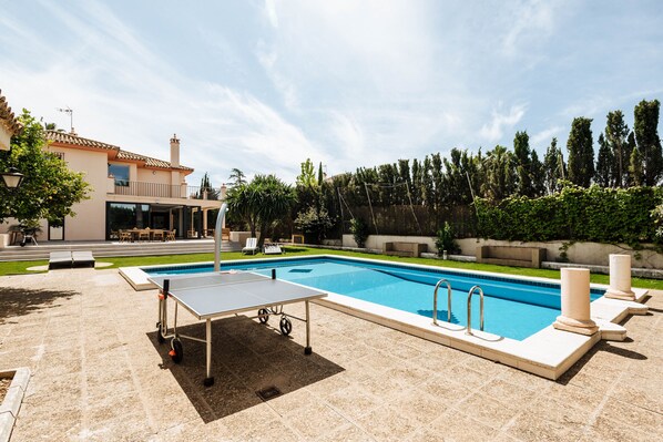 A swimming pool with a sunbathing area, a table tennis table, and a covered seating area.
