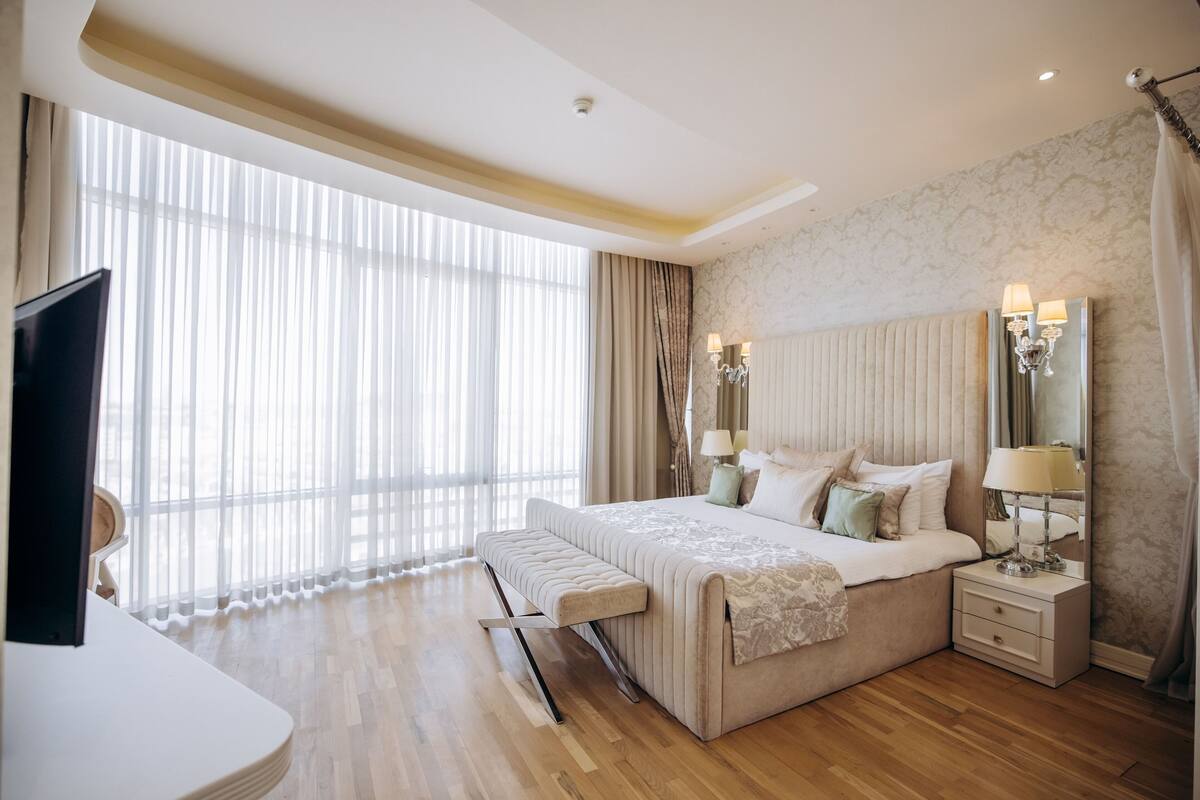 A modern hotel room with a large bed, bedside lamps, a TV, and a large window with curtains.