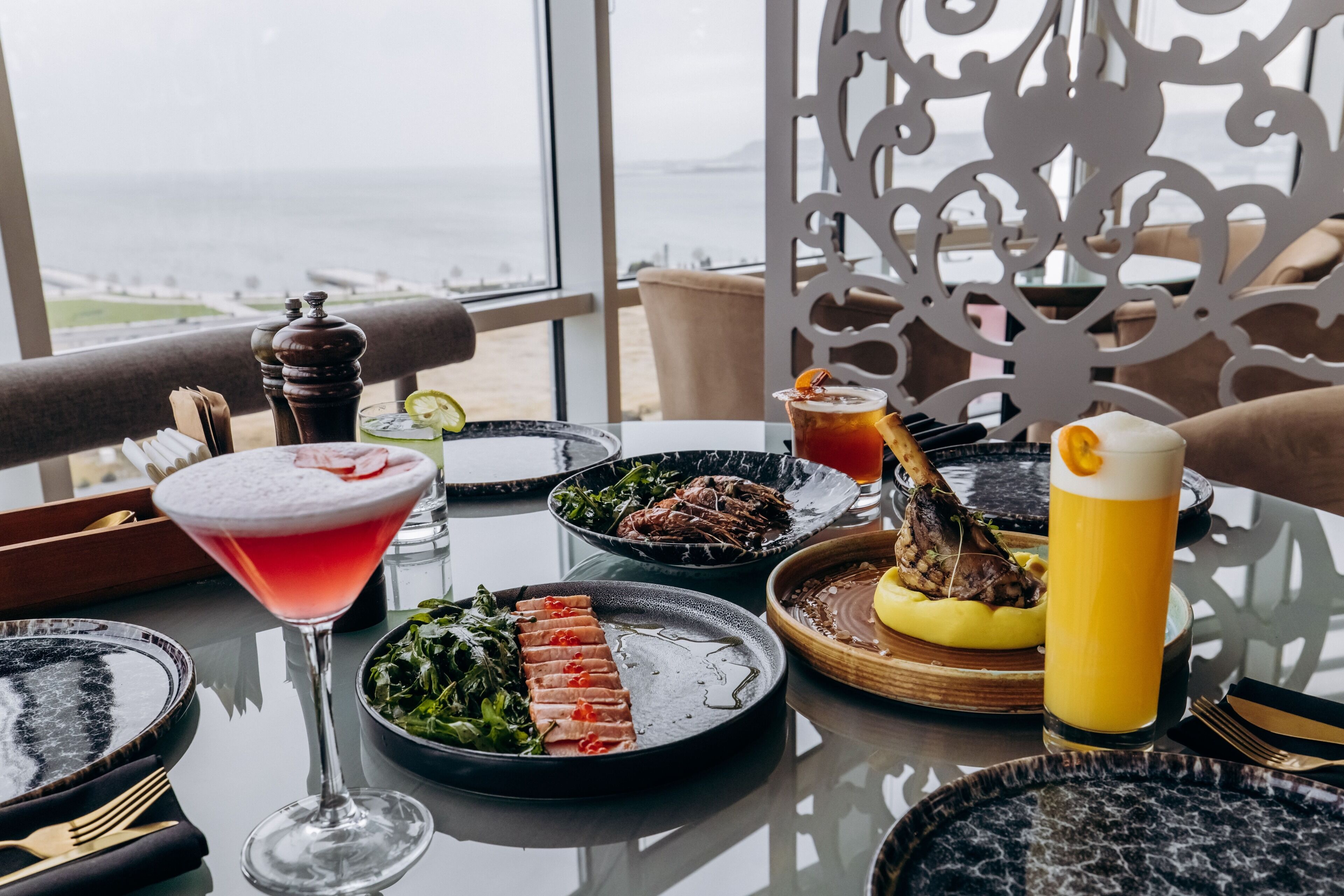 Mesa preparada para comer con diversos platos, una copa de martini y una jarra alta de cerveza, con vistas a un paisaje pintoresco.