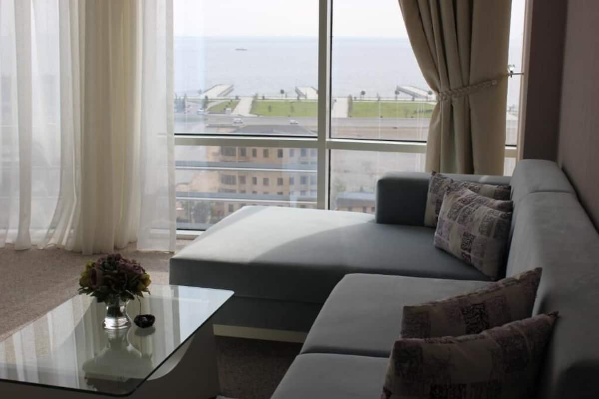 A modern living room with a grey sofa, a glass coffee table, and a view of the sea and buildings through large windows.