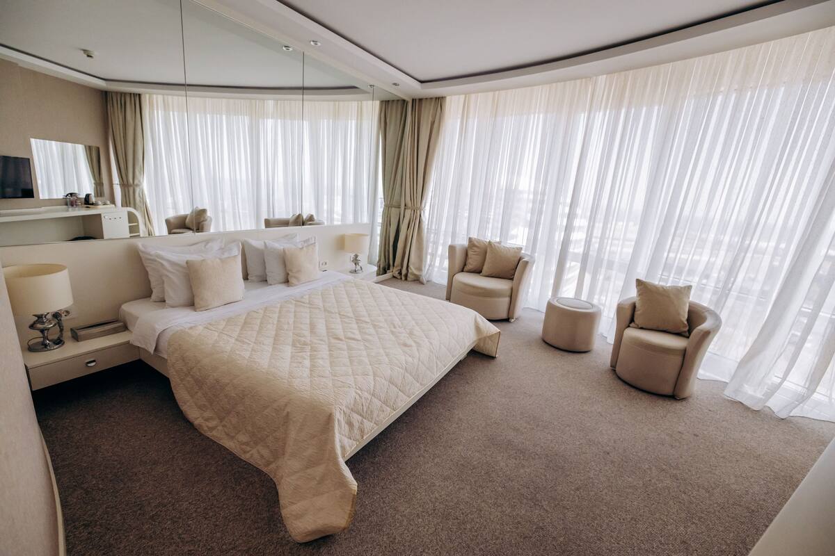 A modern hotel room with a large bed, a sitting area with a chair and ottoman, a vanity with a mirror, and a large window with sheer curtains.