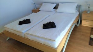 A double bed with white linens and two folded towels on each side. There are two bedside tables with lamps, and a wooden floor.