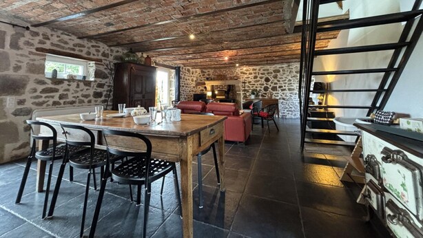 Un intérieur rustique avec des plafonds en briques apparentes, un mur en pierre, une table à manger en bois avec des chaises, un canapé en cuir rouge et un escalier menant à un niveau supérieur.