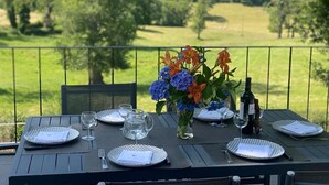 Une table dressée pour quatre personnes, avec des assiettes, des couverts et des verres, un vase rempli de fleurs et une bouteille de vin, le tout sur un balcon donnant sur un paysage verdoyant.