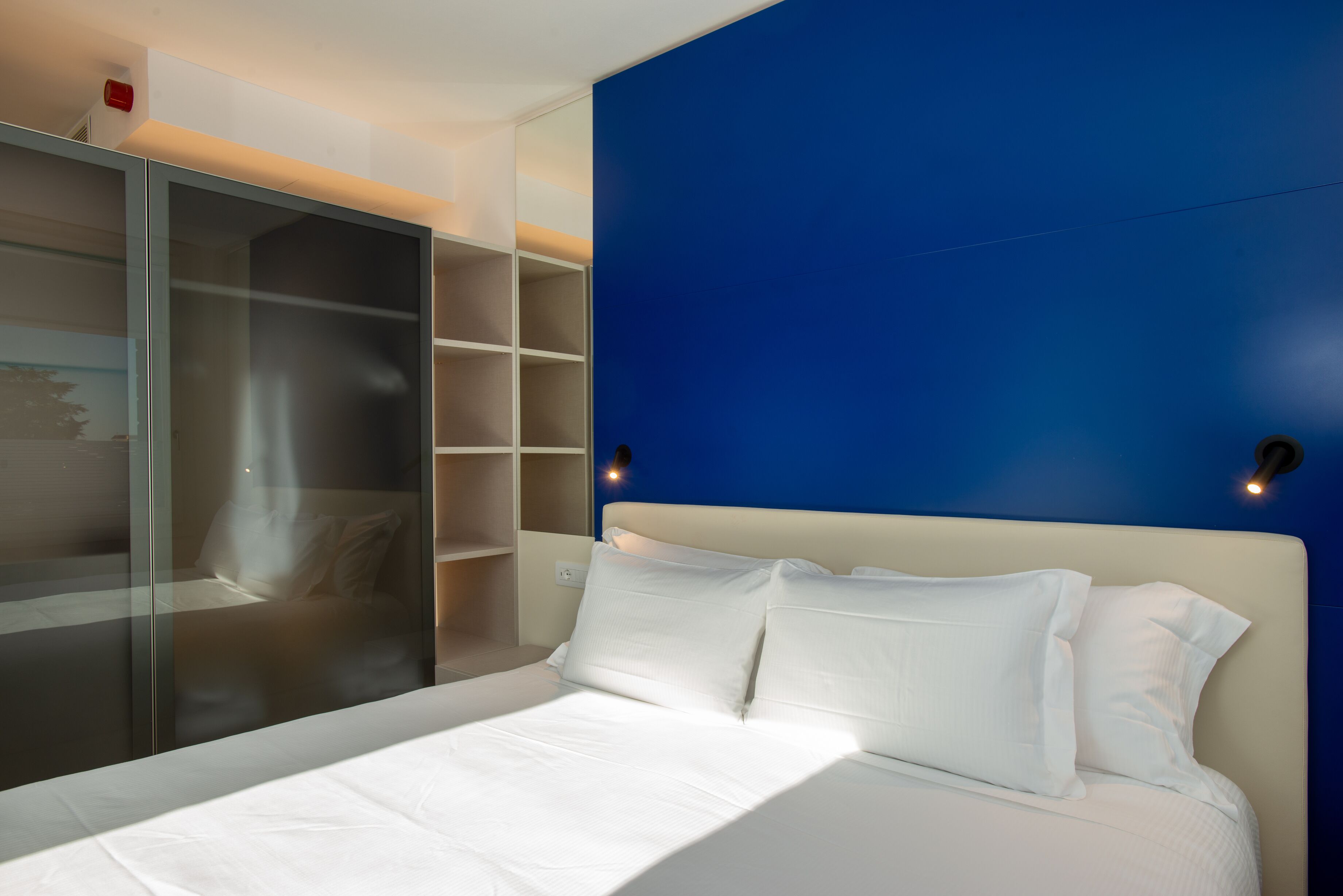 A modern bedroom with a large bed, a glass wardrobe, and a blue accent wall.