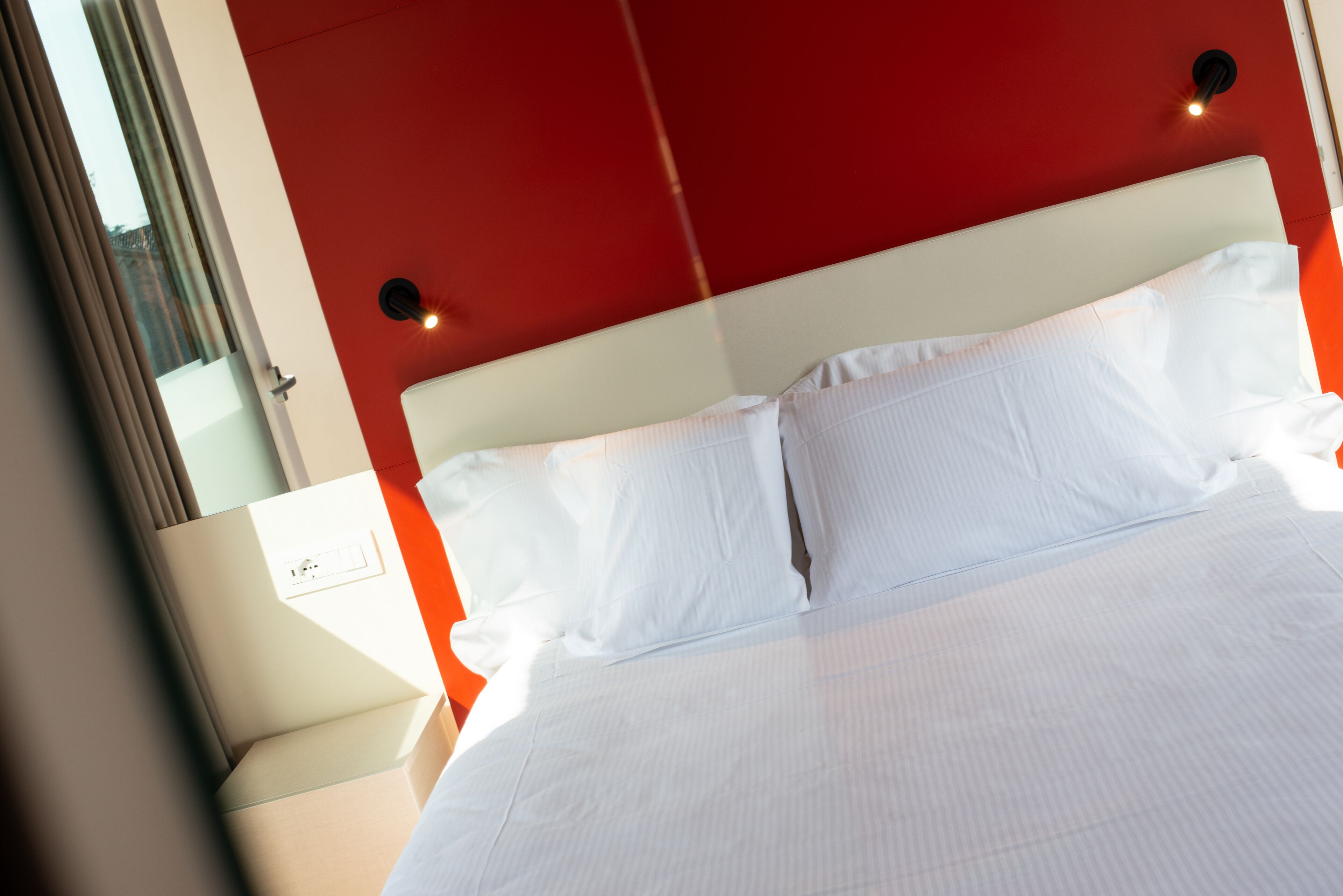 A bed with white linens, a red accent wall, and a nightstand with a power outlet.