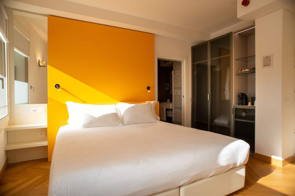 A modern hotel room with a large bed, a bright orange accent wall, and a walk-in closet.