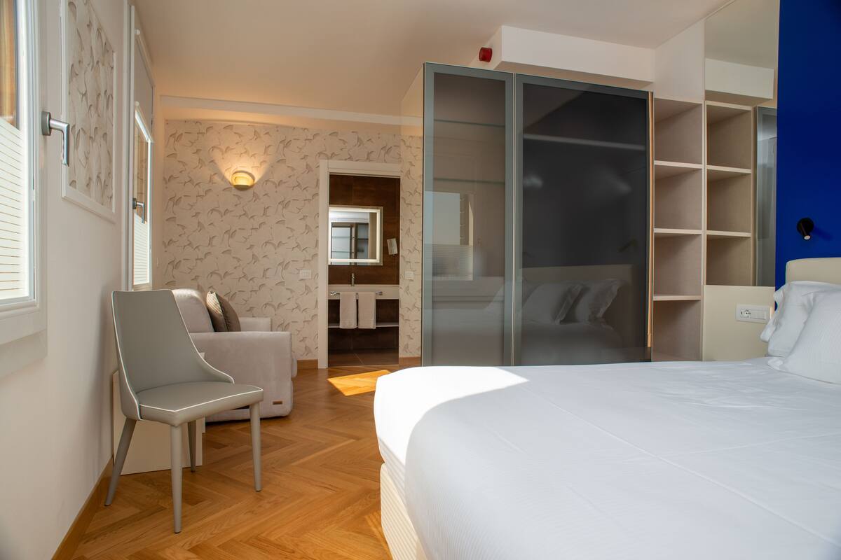 A modern hotel room with a bed, a seating area with a chair, a wardrobe, and a bathroom visible through an open door.
