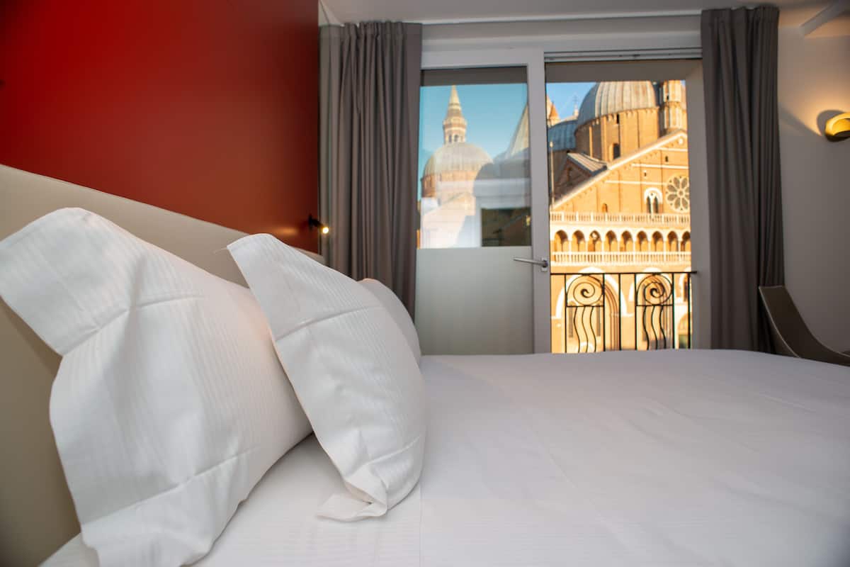 A hotel room with a large bed, a view of a historic building through a glass door, and a red wall.