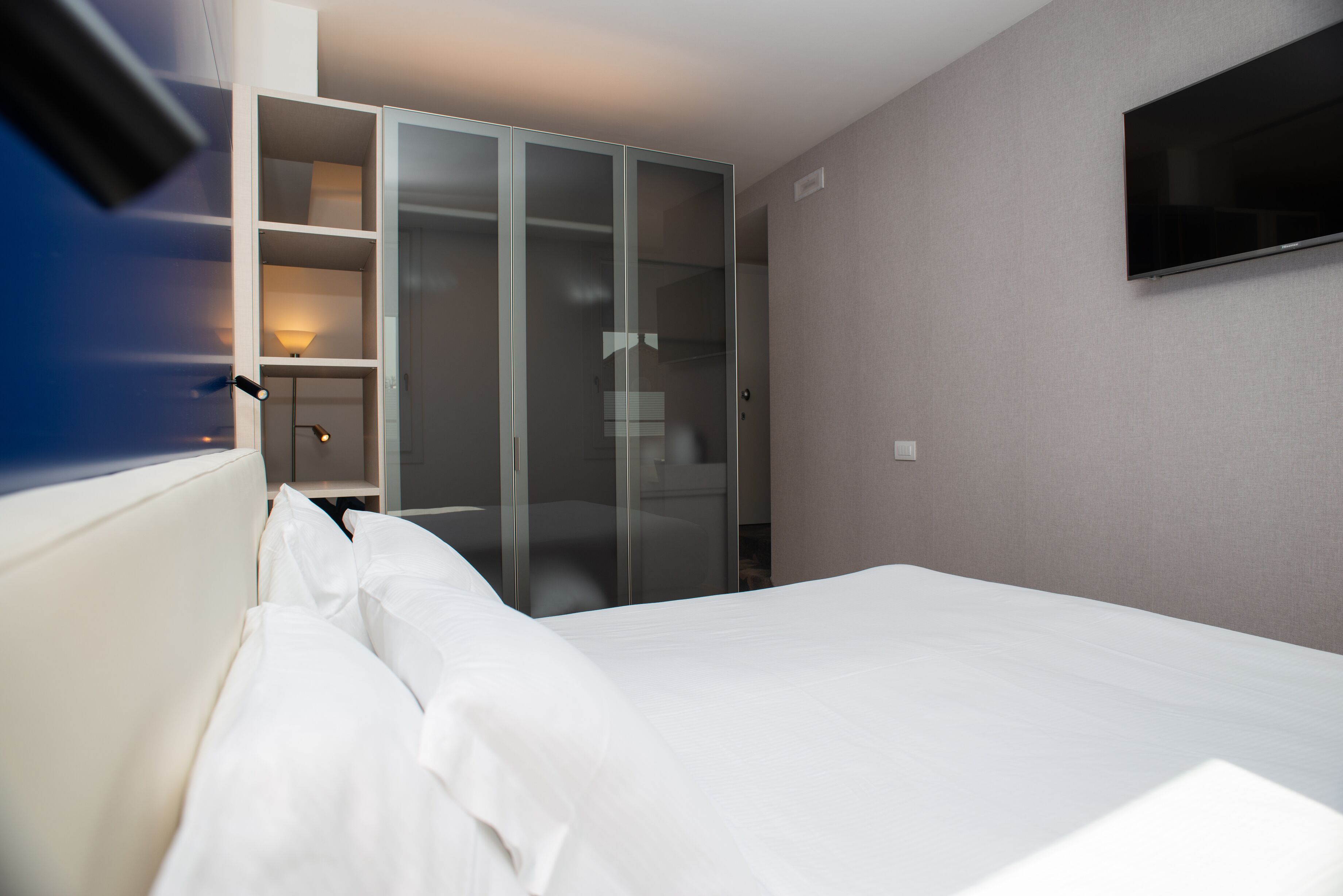 Een hotelkamer met een bed, een kledingkast, een aan de muur bevestigde televisie en een lamp op het nachtkastje.