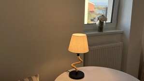 Una stanza accogliente con un tavolino rotondo, due tazze, una lampada, un cuscino, una finestra con vista e un cartello con coordinate.