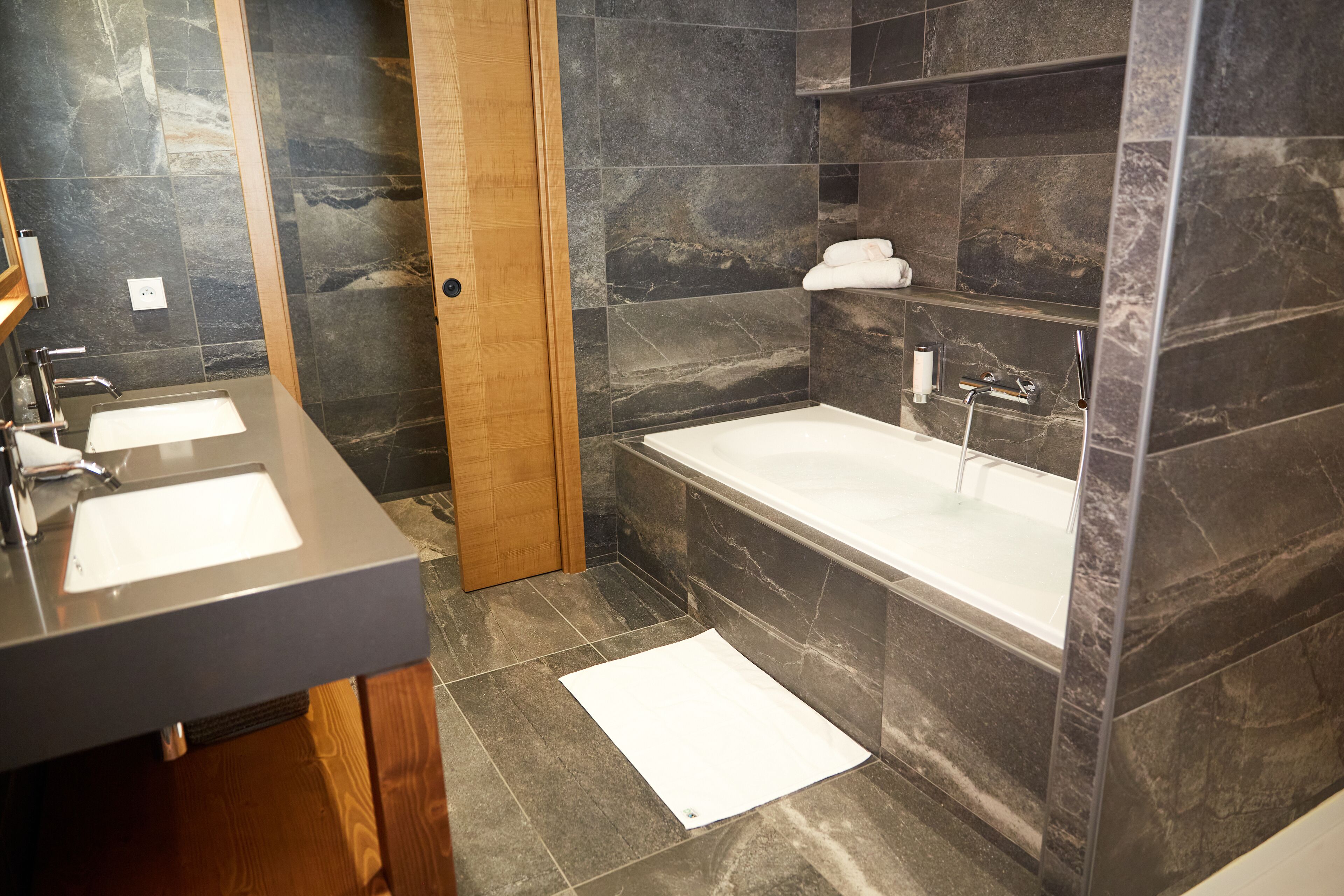 A modern bathroom with two sinks, a bathtub, and dark stone tiles.