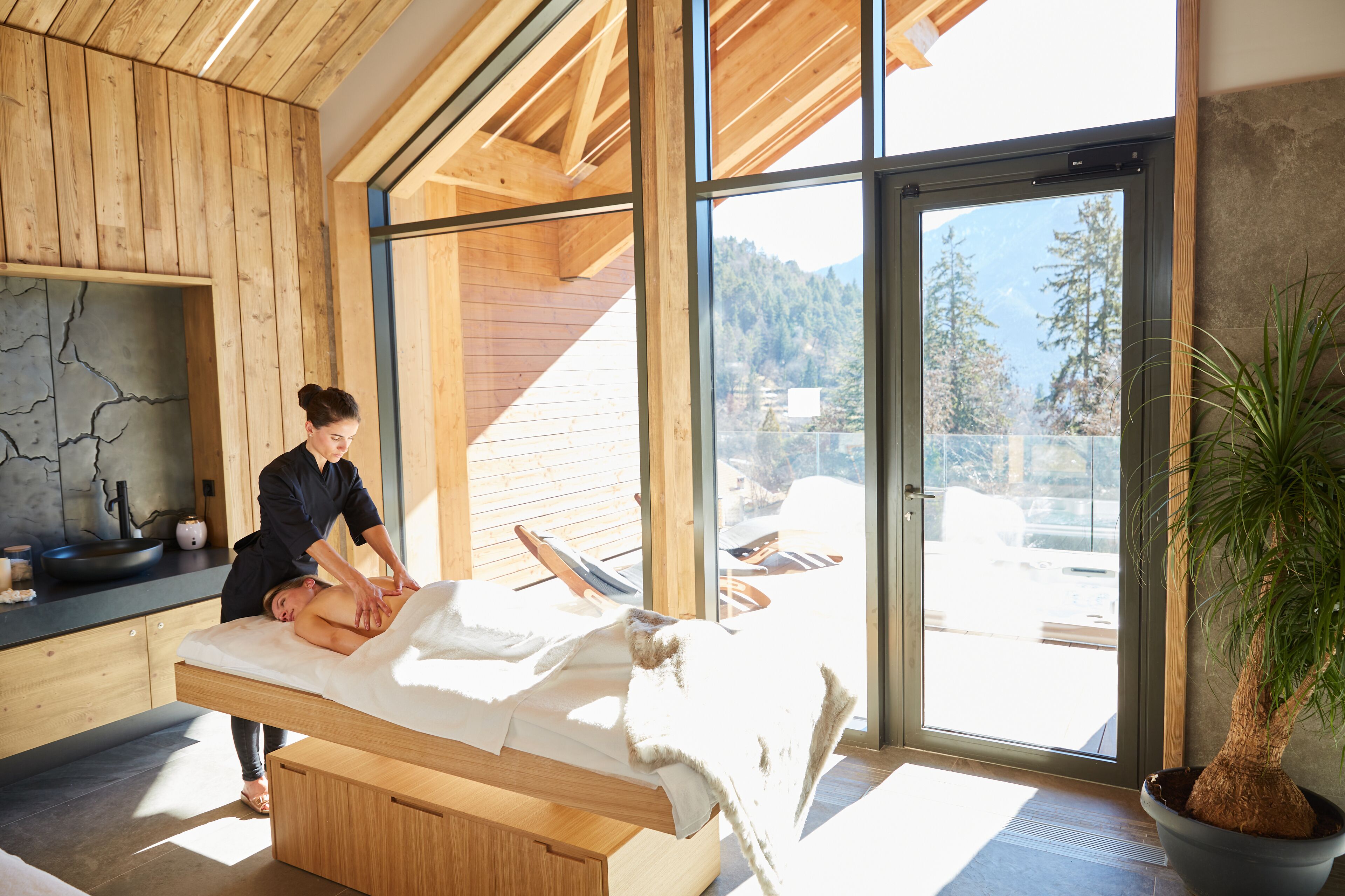 Une salle de massage avec un plafond en bois, une table de massage, une grande fenêtre donnant sur les arbres et une plante en pot.