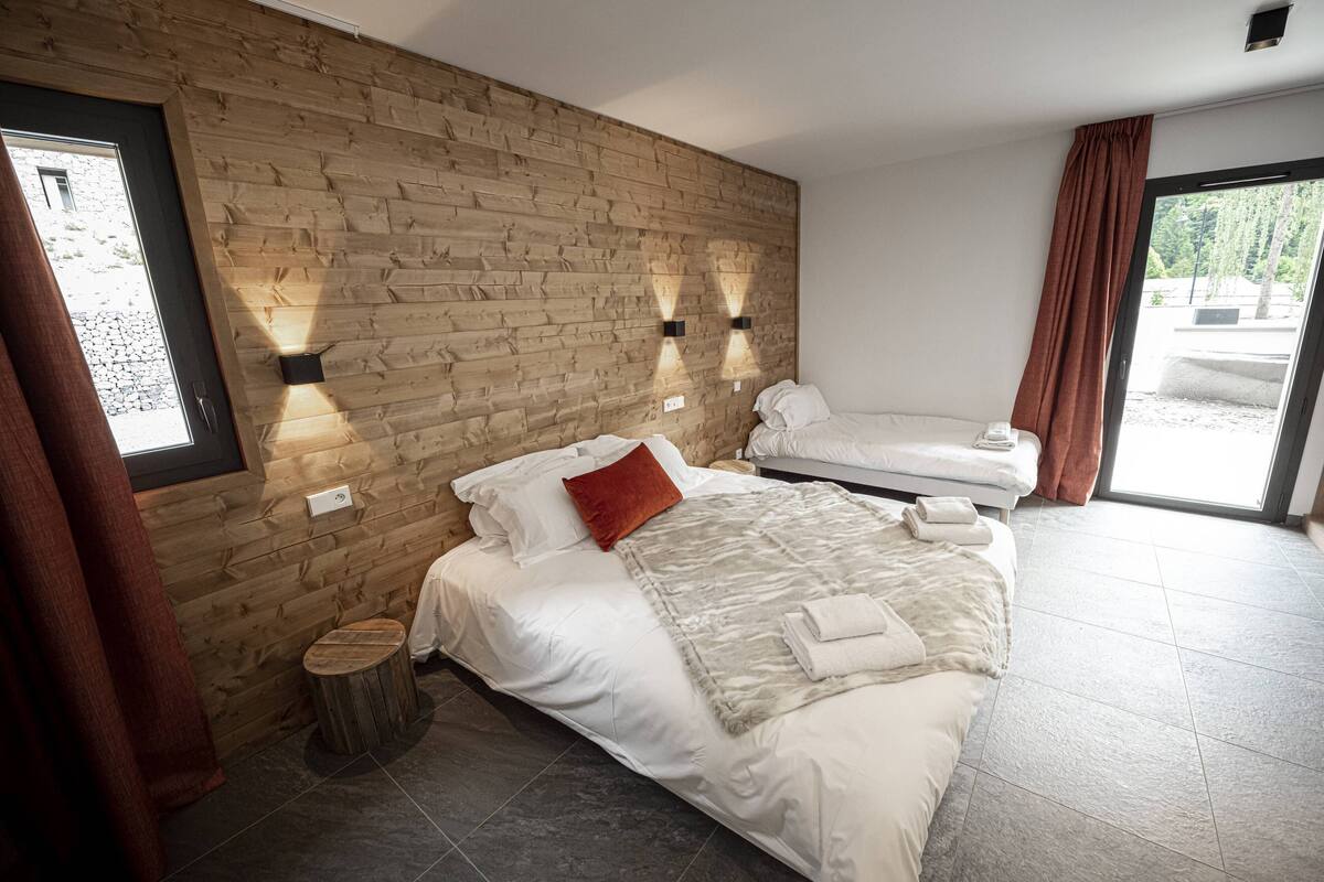 A modern bedroom with a wooden wall, two beds, a small stool, and a view through a large window.