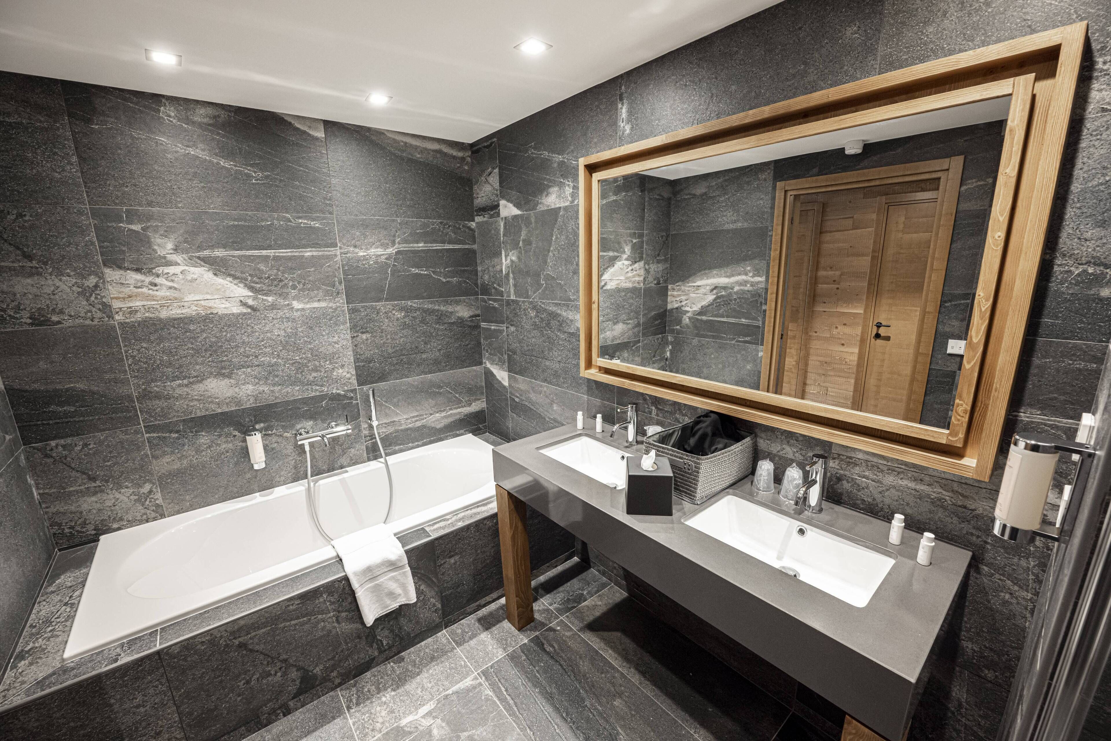 Ein modernes Badezimmer mit dunklen Marmorwänden, einem spiegeln mit Holzrahmen, zwei Waschbecken, einer Badewanne und einem Handtuchhalter.