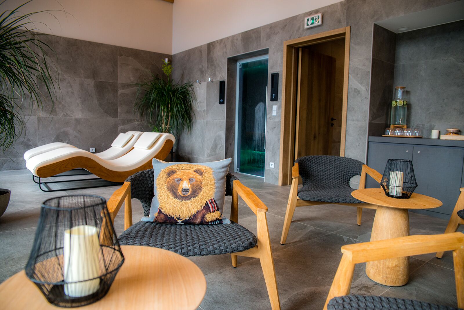 Un área de descanso moderna con un sillón de masaje, una mesa de madera con una vela y una silla con un cojín con motivos de oso.